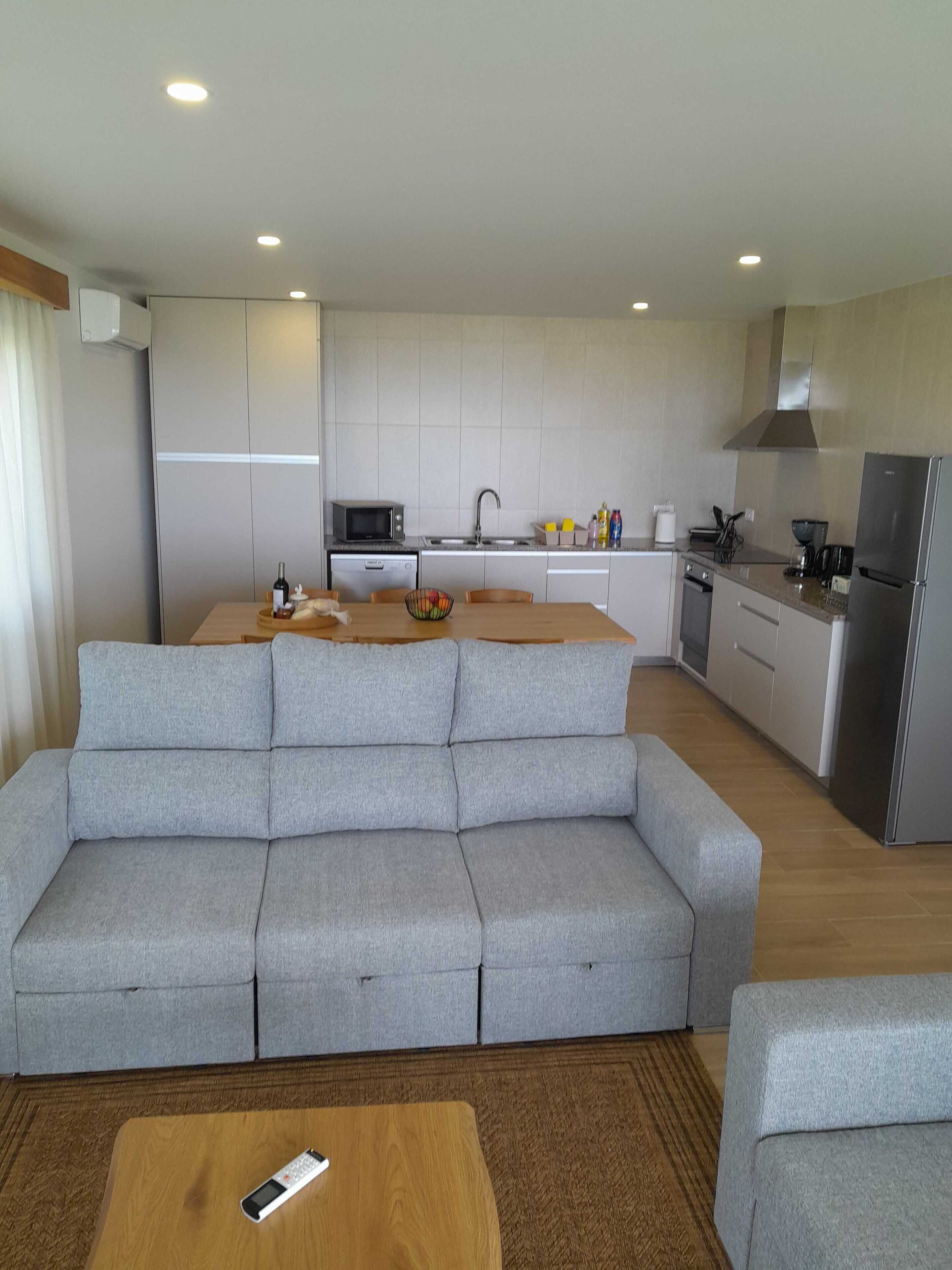 A modern living room with a grey sofa, a wooden coffee table, and a kitchen area with a microwave, sink, and refrigerator.