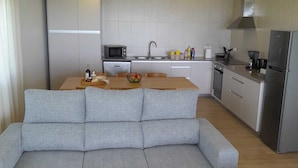 Una sala de estar moderna con un sofá gris, una mesa de centro de madera y una zona de cocina con microondas, lavaplatos y refrigerador.