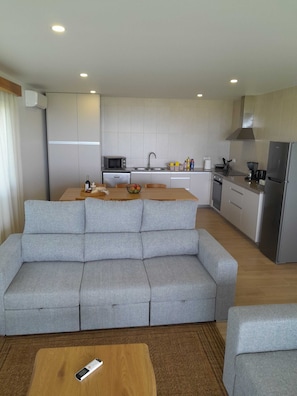 Ein modernes Wohnzimmer mit grauem Sofa, einem Holztisch und einer Küchenzeile mit Mikrowelle, Spüle und Kühlschrank.