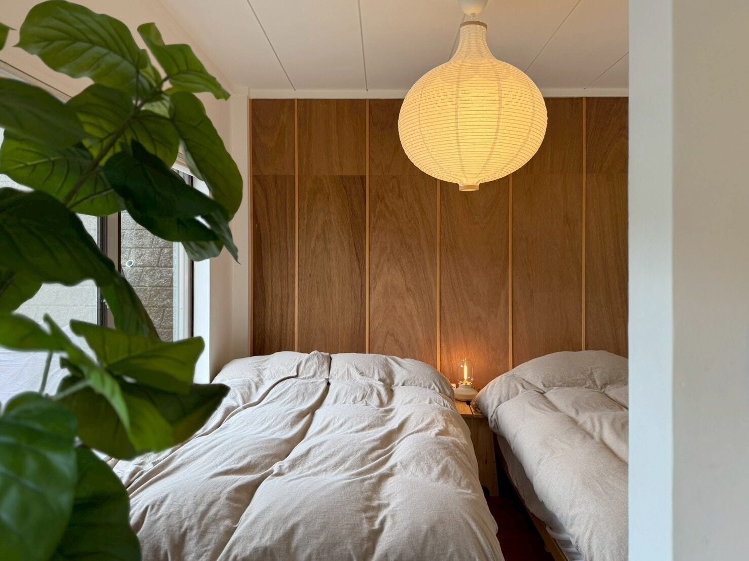 A bedroom with two beds, a wooden panel wall, a hanging light fixture, and a plant.