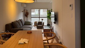 A modern living room with a sofa, a dining table, and chairs. There is a large window with a view of the outside.