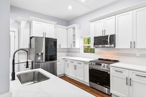 A modern kitchen with white cabinetry, stainless steel appliances, a herringbone-patterned backsplash, and a window with a view of greenery.