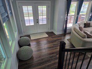 A room with a wooden floor, a white door with glass panels, a beige sofa with cushions, and a small round ottoman. - Modern 3-Bedroom Home in Downtown Raleigh - Close to EVERYTHING (Raleigh)