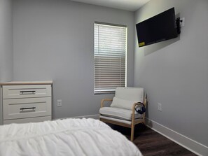 A bedroom with a bed, a bedside table, a chair, a window with blinds, and a mounted TV. - Modern 3-Bedroom Home in Downtown Raleigh - Close to EVERYTHING (Raleigh)