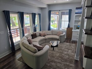 A modern living room with a curved sectional sofa, a glass coffee table, and a view of the outdoors through large windows. - Modern 3-Bedroom Home in Downtown Raleigh - Close to EVERYTHING (Raleigh)