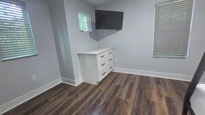 Ein Zimmer mit Holzboden, einer weißen Kommode mit Schubladen, einem an der Wand befestigten Flachbildfernseher und zwei Fenstern mit Jalousien.