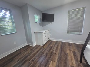 Ein Zimmer mit Holzboden, einer weißen Kommode mit Schubladen, einem an der Wand befestigten Flachbildfernseher und zwei Fenstern mit Jalousien.