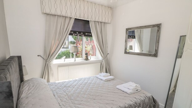 A bedroom with a bed, a window with curtains, a mirror, and a radiator. - Beachway (Holywell)