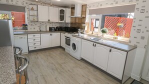 A modern kitchen with white cabinetry, a built-in oven, a washing machine, and a window with a view of a red fence and greenery. - Beachway (Holywell)