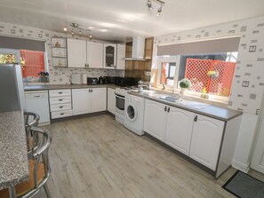 A modern kitchen with white cabinetry, a built-in oven, a washing machine, and a window with a view of a red fence and greenery. - Beachway (Holywell)