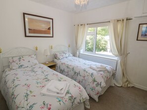 Ein Schlafzimmer mit zwei Betten, einem Fenster mit Vorhängen, einem Nachttisch mit Lampe und einem gerahmten Bild an der Wand.