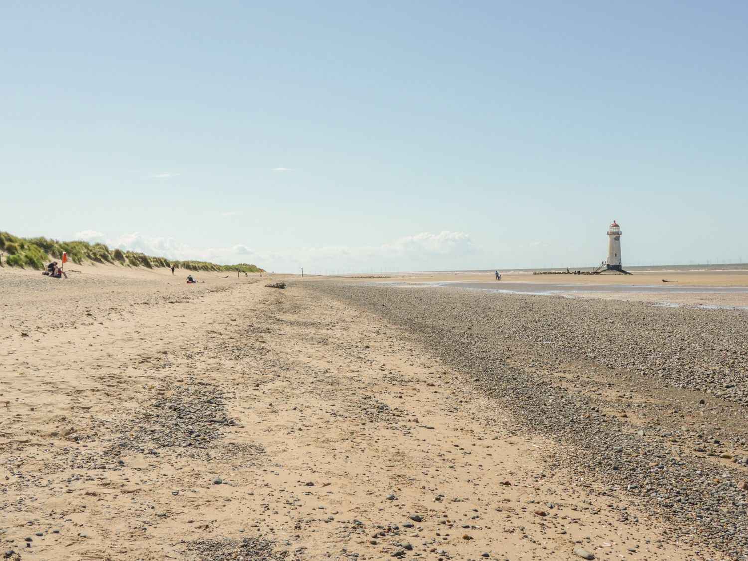 Una playa con un faro a lo lejos, terreno arenoso y algunas personas.