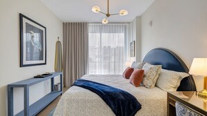 Habitación de hotel con cama, mesita de noche, lámpara, cortinas e imagen enmarcada en la pared.