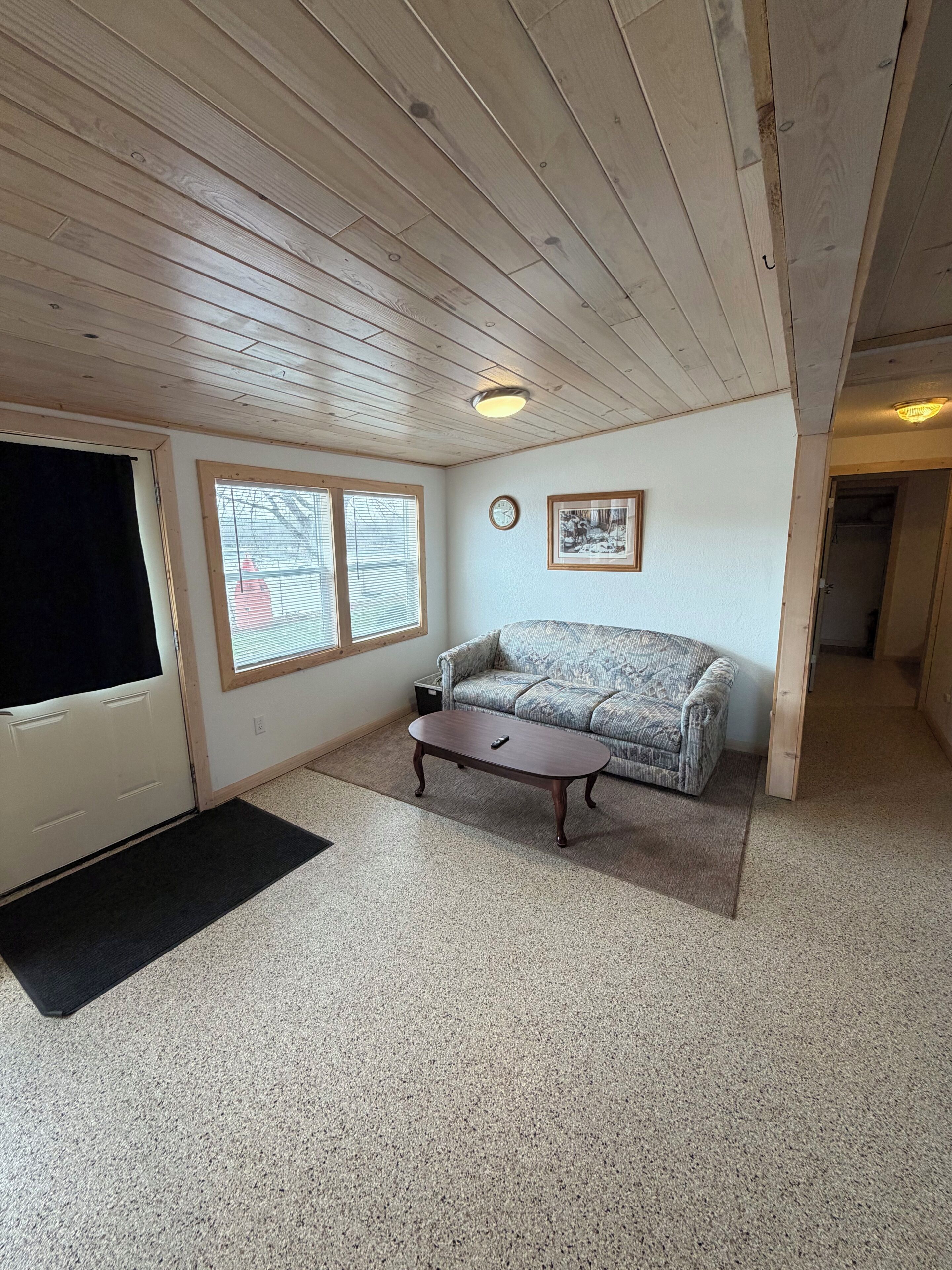 Ein Zimmer mit Holzboden, einer Couch, einem Couchtisch, einem Fenster mit Jalousien, einer Tür und einem gerahmten Bild an der Wand.