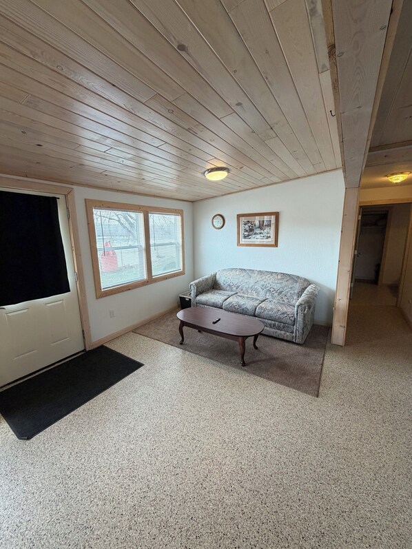 Ein Zimmer mit Holzboden, einer Couch, einem Couchtisch, einem Fenster mit Jalousien, einer Tür und einem gerahmten Bild an der Wand.