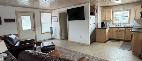 A spacious living area with a leather sofa, a television mounted on the wall, and a kitchen area with wooden cabinets and a window.