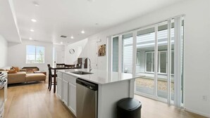 A modern kitchen with a central island, a dining area with a table and chairs, a sofa, and a large sliding glass door leading to an outdoor area. - Aurora Modern Retreat (Aurora)