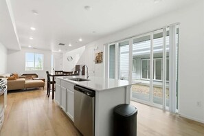 Eine moderne Küche mit Kochinsel, Essbereich mit Tisch und Stühlen, Sofa und große Schiebeglastür zu einem Außenbereich.