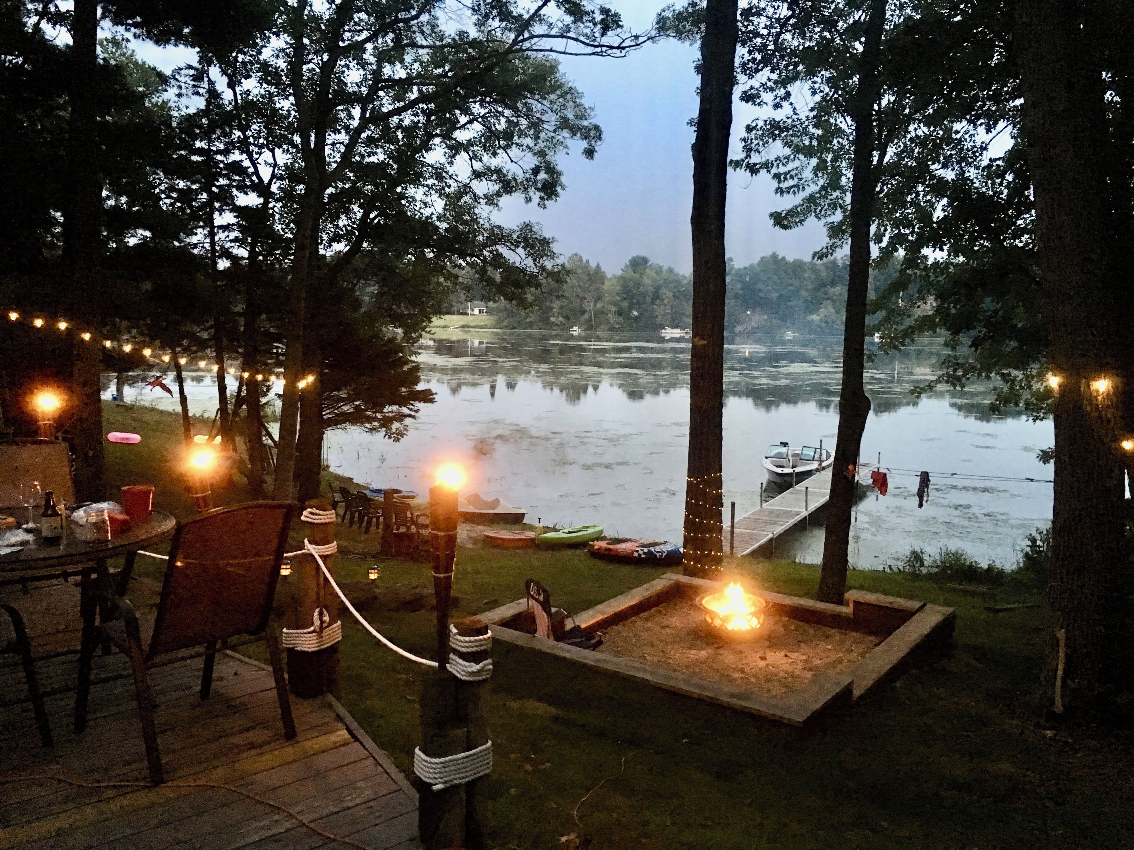 An outdoor dining area with a view of a lake, surrounded by trees, and featuring string lights.