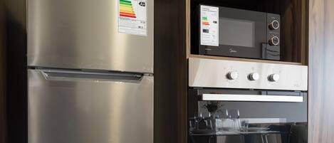 Uma geladeira de aço inoxidável com etiqueta de eficiência energética, um micro-ondas e um forno embutido em uma cozinha com armários de madeira.