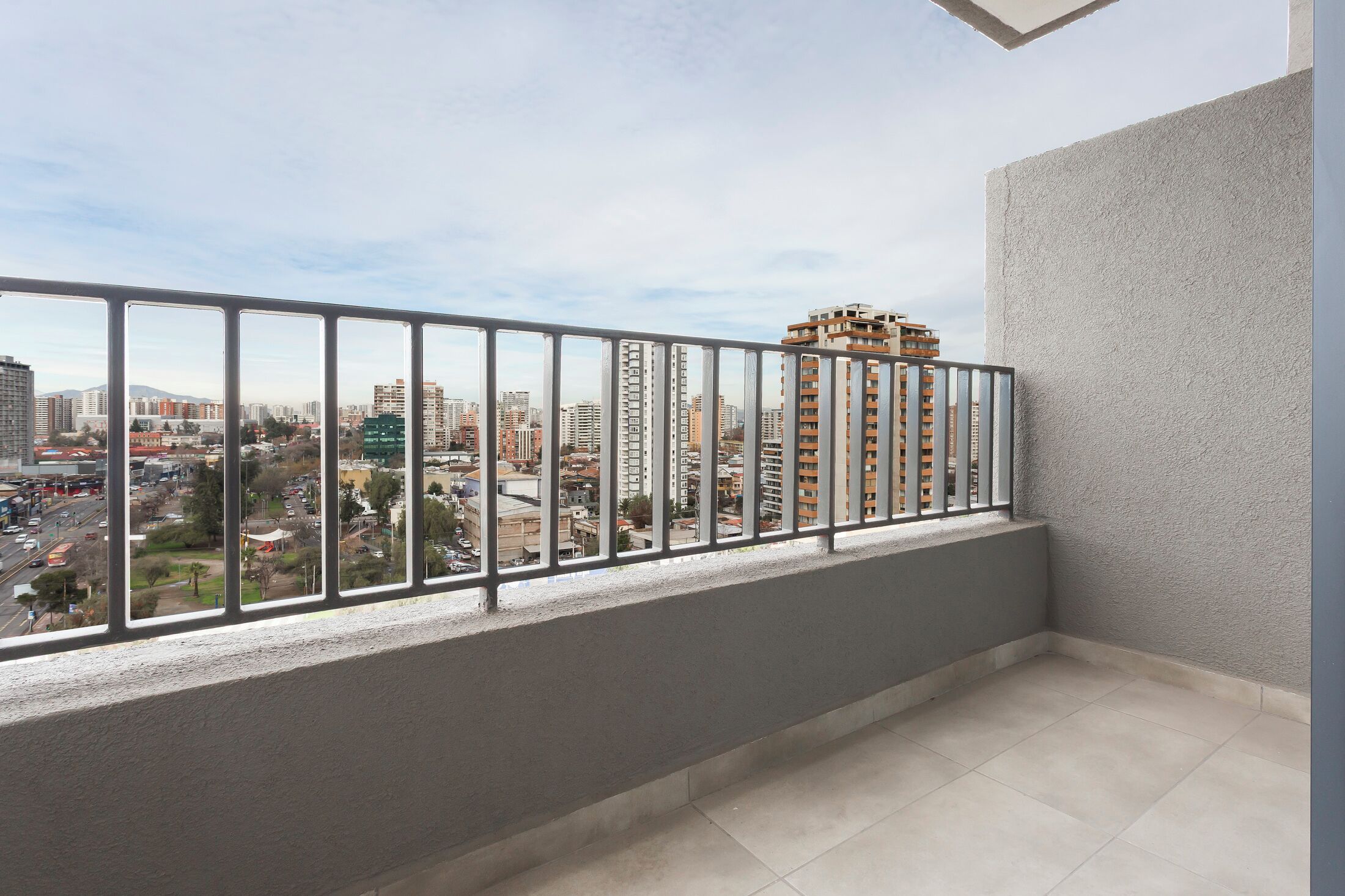 Un balcon doté d’une rambarde en métal, d’un sol carrelé et offrant une vue sur un paysage urbain avec des bâtiments et une route.