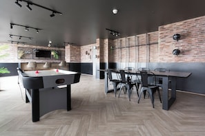 A modern indoor space with a foosball table, a long dining table with chairs, and a brick wall with a mounted television.