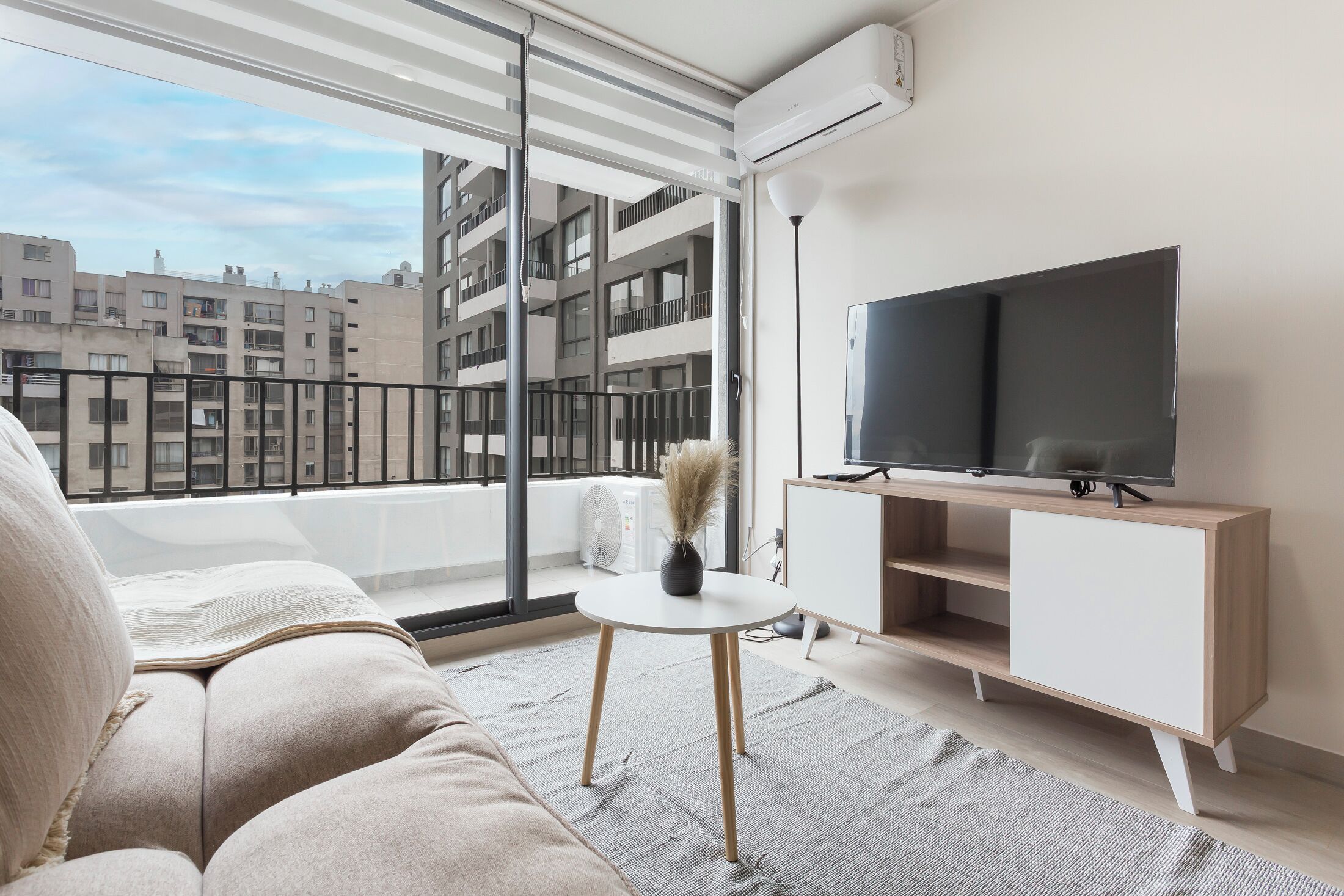 Un salon moderne avec un canapé, une petite table, un téléviseur et un balcon offrant une vue sur les immeubles.