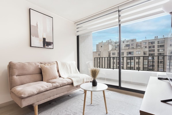Una sala de estar moderna con un sofá, una mesita redonda con un florero y un ventanal con vista a la ciudad.