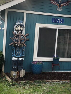 Une maison dont le cadre de la fenêtre porte le numéro « 15716 », une sculpture en bois représentant une roue de navire, et une plante en pot placée à côté de la fenêtre.