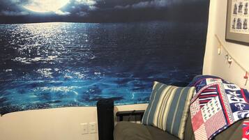 Una habitación con un tapiz grande que muestra una escena oceánica a la luz de la luna, un sofá gris con cojines estampados y una mesita con una lámpara.