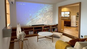 Ett modernt vardagsrum med en stor skärm som visar ett naturbild, ett träbord, en soffa och ett köksområde som syns genom en öppen dörröppning.