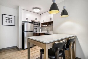 A modern kitchen with white cabinets, a stainless steel refrigerator, a microwave, and a wooden dining table with chairs.