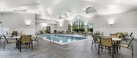 Un espace piscine intérieur moderne, doté d’un plafond au design géométrique, entouré de tables et de chaises.