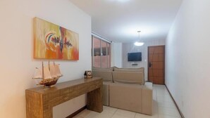 A modern living room with a wooden console table, a beige sofa, a large abstract painting, and a television mounted on the wall. - 2-bedroom Apt 150m From the Beach Tib0103 (Guarapari)