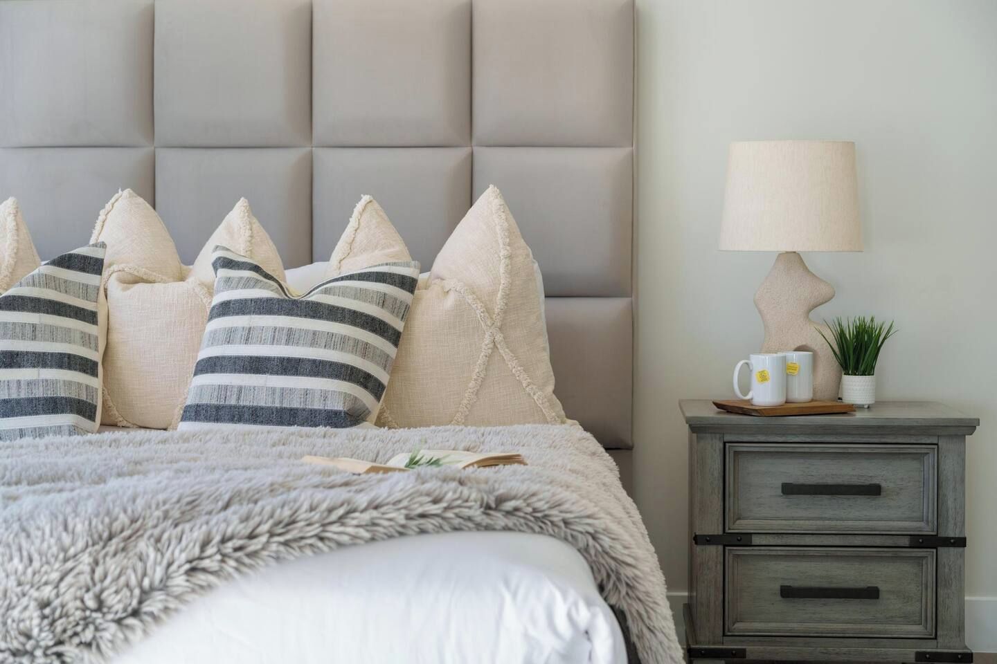 A bedroom with a tufted headboard, striped pillows, a fluffy throw, a bedside lamp, a small plant, and a nightstand with drawers.