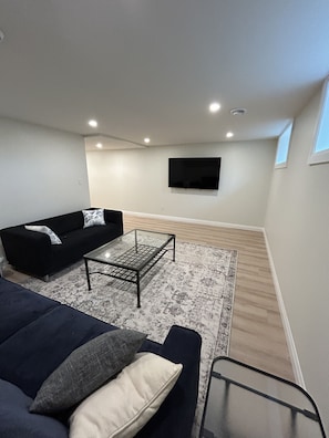 A living room with a black sofa, a glass coffee table, a patterned rug, and a flat-screen TV mounted on the wall.