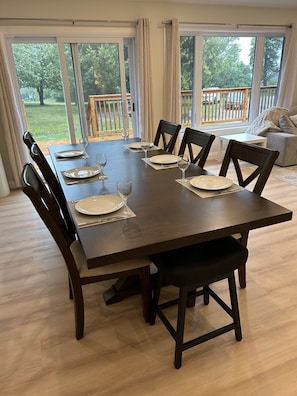 A dining area with a large table set for six, surrounded by chairs, with a view of a wooden deck and greenery outside.