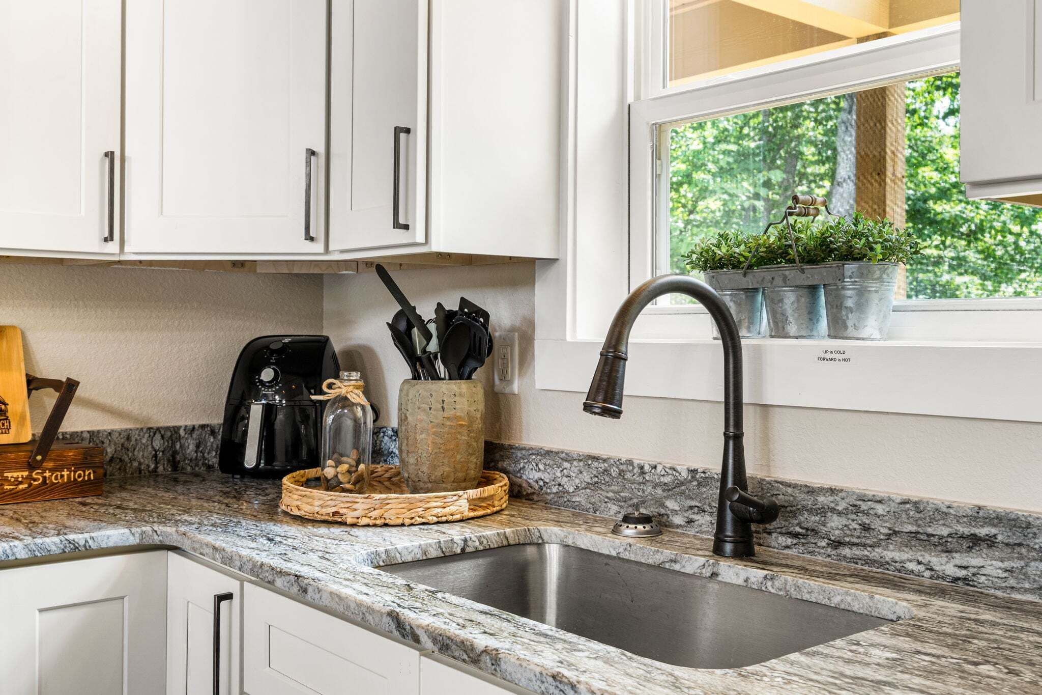 A kitchen with white cabinets, a granite countertop, a stainless steel sink, a coffee maker, and a potted plant on the windowsill.