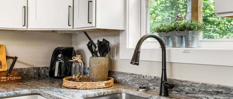 A kitchen with white cabinets, a granite countertop, a stainless steel sink, a coffee maker, and a potted plant on the windowsill.
