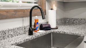 Una cucina con un lavello nero, una finestra con persiane, una bottiglia di detersivo Ajax e un asciugamano.