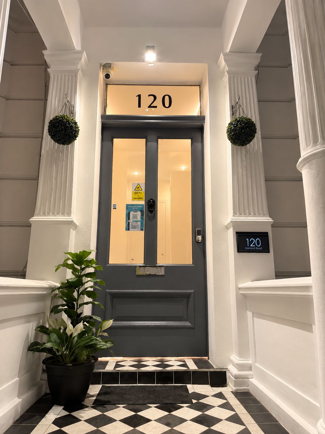 Een hotelentree met een grijze deur, nummer 120, een waarschuwingsbord voor bewakingscamera's en een bloempot aan de linkerkant.