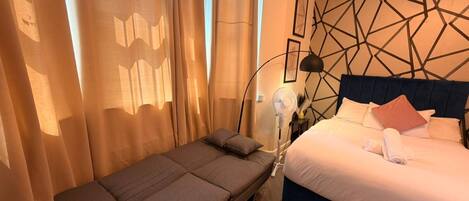 A hotel room with a bed, a sofa, a standing fan, and curtains.