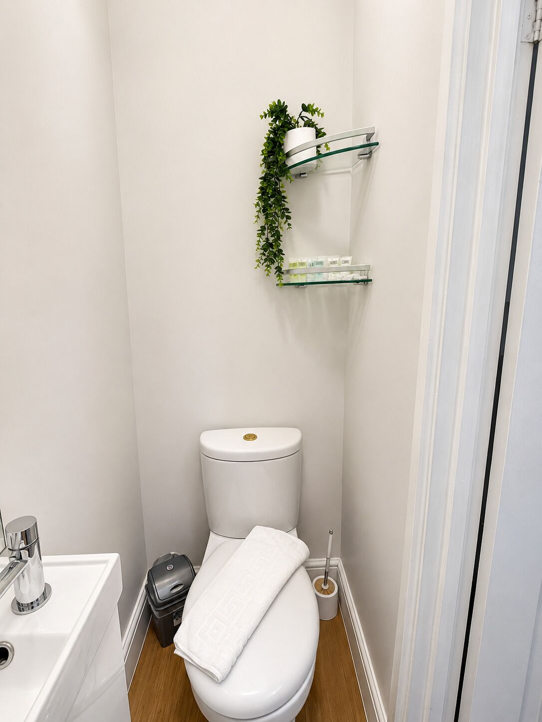 Une salle de bain avec des toilettes blanches, un lavabo et une poubelle. Deux étagères en verre sont fixées au mur : l’une supporte un pot de fleurs et l’autre contient des articles de toilette.