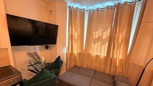 A compact room with a flat-screen TV mounted on the wall, a glass table with a vase and a remote, a green chair, a sofa, and sheer curtains. - Cozy Flats Near Olympia and Westfield (London)