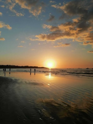 Une scène de plage au coucher du soleil, avec des eaux calmes et les silhouettes de personnes sur le rivage.