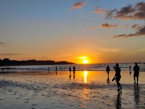 Eine Strandansicht mit Menschen, die gehen und stehen, einem Sonnenuntergang im Hintergrund und ruhiger See.