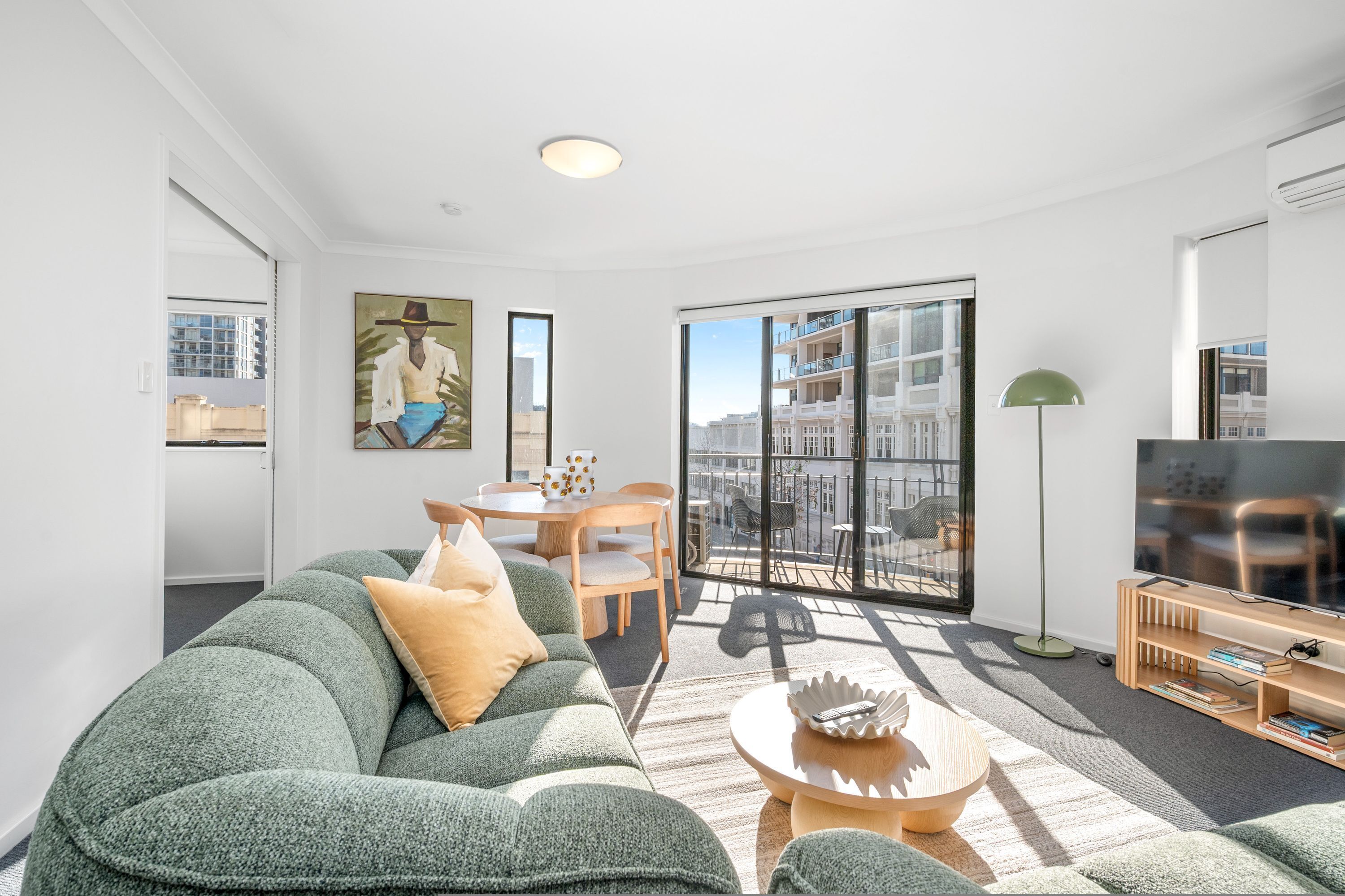 A modern living room with a sofa, a coffee table, a painting, and a balcony with a view of buildings.