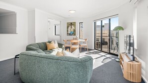 A modern living room with a green sofa, a dining table with chairs, and a large window with a view of the city. - Rest and Roam - A Executive Sepal Stay (Perth)