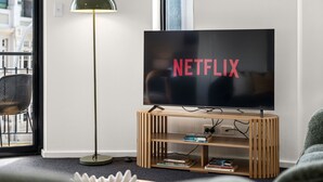 A modern living room with a television displaying the Netflix logo, a floor lamp, a bookshelf with books, and a view of a building outside. - Rest and Roam - A Executive Sepal Stay (Perth)