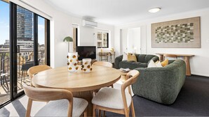 A modern living room with a round wooden table, green sofa, and a city view through large windows. - Rest and Roam - A Executive Sepal Stay (Perth)